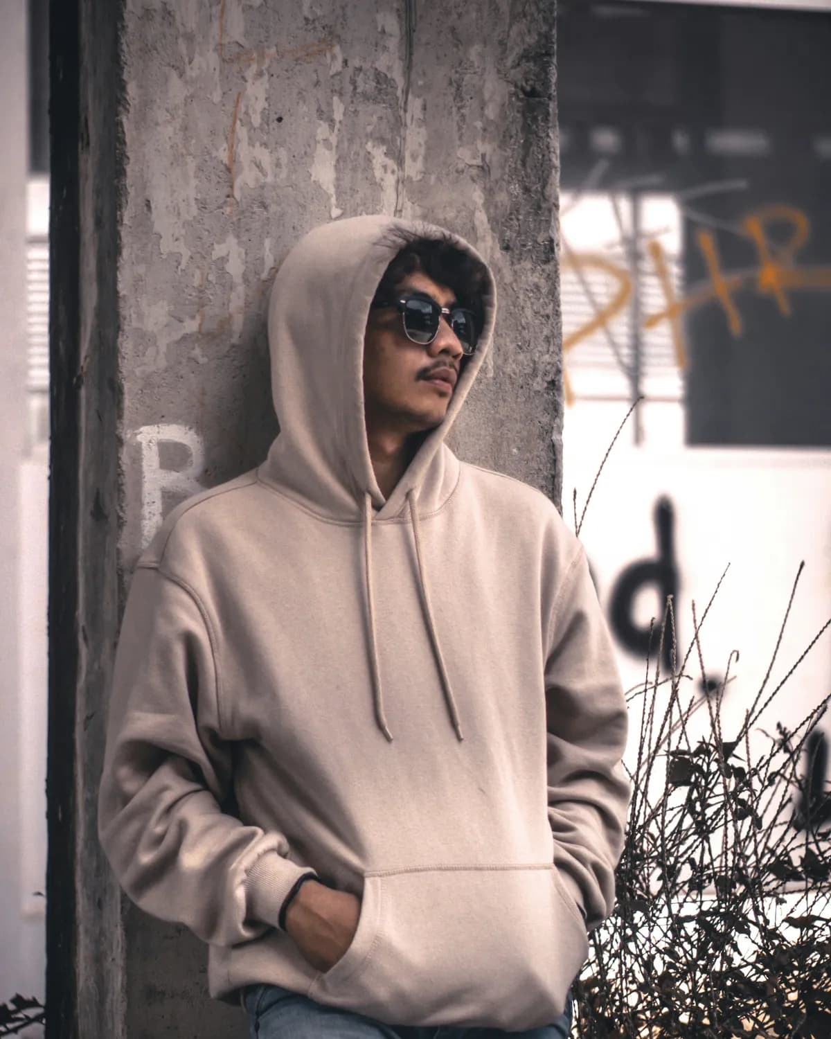 A man in a plain beige hoodie leaning on a concrete pillar