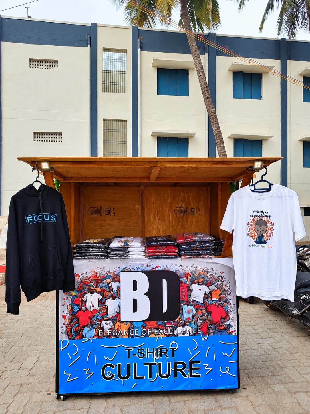 The first BlingDuds stall — a wooden cart under a banner reading BD, Elegance of Excellence, T-shirt Culture, with folded tees stacked on the counter and a hoodie and a printed tee hanging either side.